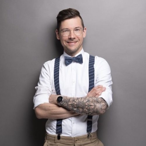 Ein weißes Mann mit weißem Hemd, blauer Fliege und Hosenträger. Die Arme sind gekreuzt und der linke Arm ist tätowier. Der Mann trägt eine Brille, lacht und hat kurze braune Haare.