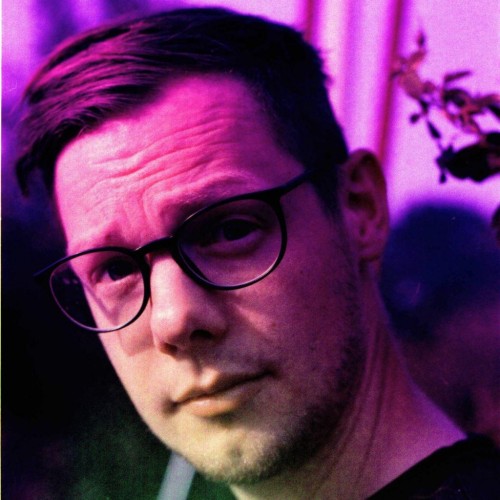 Portrait aufgenommen auf einem analogen Effektfilm mit einem horizontalen Farbverlauf der im größten Teil lila ist und im unteren Teil des Bildes zu natürlichen Farben verläuft. Die Person hält ihren kopf schräg, guckt zur Seite direkt in die Kamera. Die Person trägt eine Brille